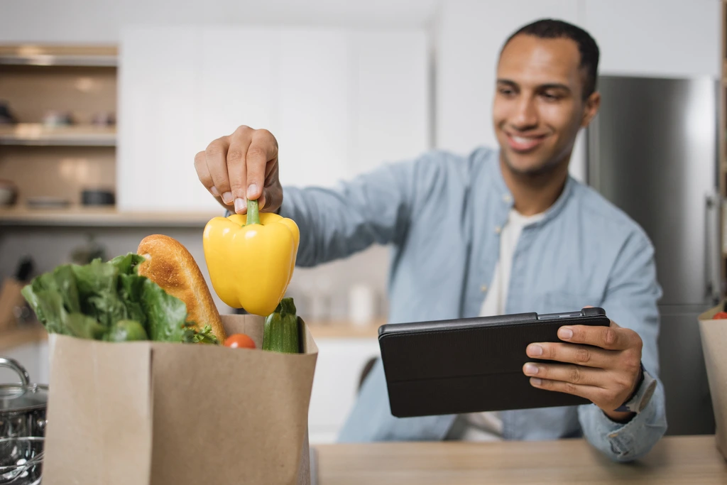 Leo Grant, the author of Dishicious, smiling as he selects a fresh yellow bell pepper from a grocery bag while holding a tablet.