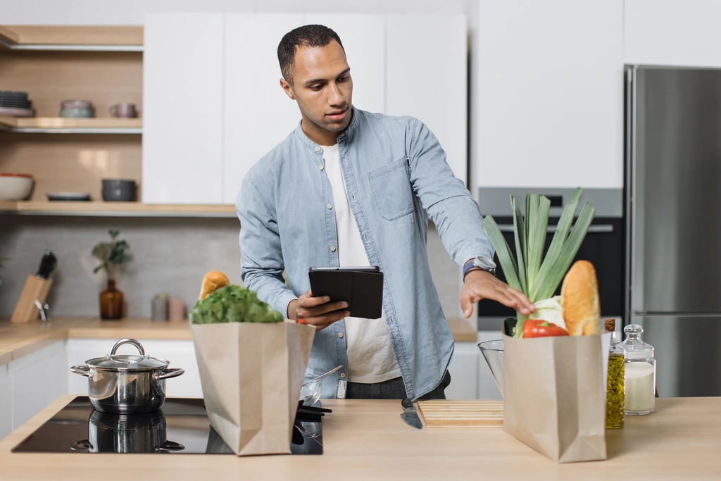 Leo Grant, the founder of Dishicious, standing in his kitchen, looking at a tablet while unpacking grocery bags filled with fresh vegetables and bread.
