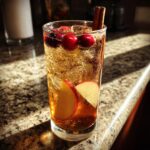 A tall glass filled with Apple Cider Long Island Iced Tea, ice, cranberries, apple slices, and a cinnamon stick.