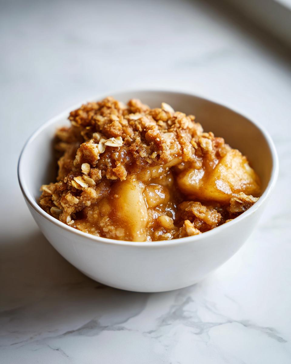 Close-up of a white bowl filled with warm, gooey apple crumble topped with oats and spices.