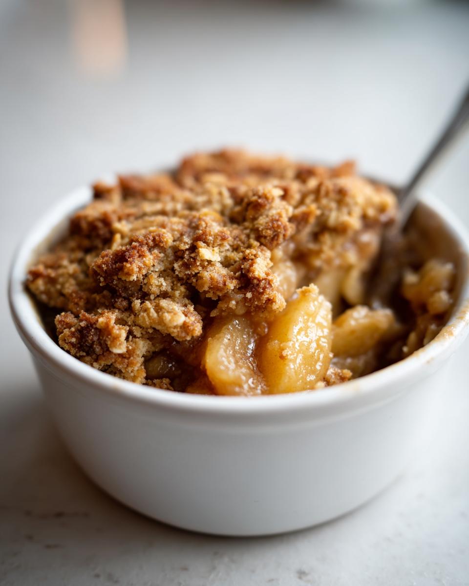 Close-up of a warm apple crumble in a white bowl, with a spoon ready to dig in.