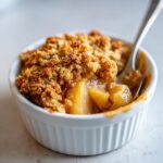 Close-up of a warm, comforting apple crumble in a white ramekin, with a spoon digging in.
