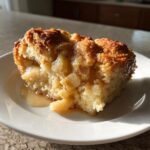 A close-up of a moist slice of apple pie dump cake on a white plate, showing tender apple chunks and a golden crumb topping.