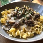 A close-up of a bowl filled with creamy Beef Stroganoff served over wide egg noodles, garnished with fresh parsley.