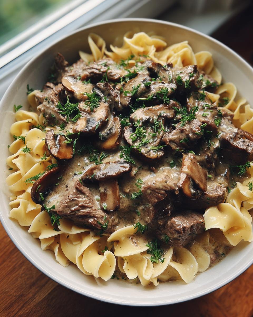 A close-up of a bowl of creamy Beef Stroganoff served over wide egg noodles, topped with mushrooms and fresh dill.
