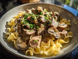 A close-up of a bowl of beef stroganoff served over egg noodles, topped with mushrooms and fresh parsley.