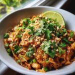 A vibrant bowl of Chicken Pad Thai topped with fresh cilantro, chopped peanuts, and a lime wedge.