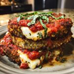 A close-up of a stack of crispy Eggplant Parmesan, topped with marinara sauce and fresh basil, with melted mozzarella oozing out.