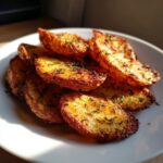 A close-up of a pile of golden brown crispy potato wedges seasoned with herbs on a white plate.