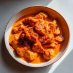 A close-up overhead view of tender pieces of crockpot butter chicken in a rich, creamy orange sauce, served in a rustic bowl.
