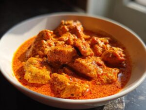 A close-up of a white bowl filled with tender pieces of crockpot butter chicken in a rich, creamy orange sauce.