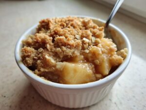 Close-up of a warm apple crumble in a white ramekin, topped with crunchy crumble and a spoon.