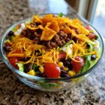 A close-up of a colorful Dorito Taco Salad in a glass bowl, featuring ground beef, cheese, tomatoes, corn, beans, and Dorito chips.
