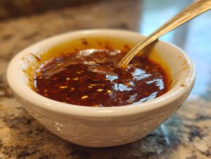 A close-up of a white bowl filled with rich, dark Dumpling Sauce, speckled with sesame seeds, and a spoon resting inside.
