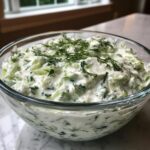 A close-up of a clear glass bowl filled with creamy Tzatziki sauce, featuring chopped cucumbers and fresh dill.