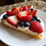A slice of fruit pizza sugar cookie topped with cream cheese frosting, strawberries, and blueberries.