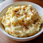 A close-up of creamy garlic parmesan mashed potatoes topped with crispy bits and herbs in a white bowl.
