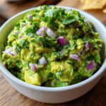 A close-up of a white bowl filled with chunky, vibrant green homemade guacamole, topped with red onion and cilantro.