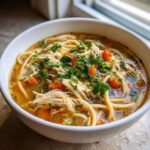 A comforting bowl of Italian penicillin soup, featuring shredded chicken, pasta noodles, diced carrots, and fresh parsley.