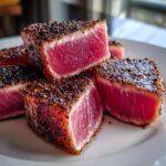 Close-up of perfectly seared Tuna Steaks, cut into bite-sized pieces, showcasing a vibrant pink interior and a dark pepper crust.