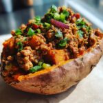A baked sweet potato split open and filled with savory ground meat mixture, topped with chopped green onions and red onion.