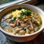 A close-up of a steaming bowl of white chicken chili, topped with shredded chicken, white beans, cheese, and cilantro.