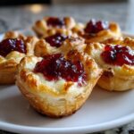 Close-up of warm, flaky puff pastry brie bites topped with melted brie and bright red jam.