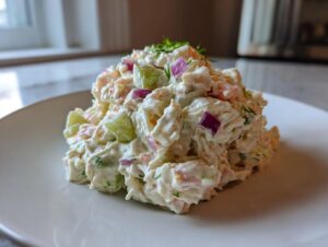A mound of creamy crab salad mixed with diced red onion and cucumber, garnished with dill, served on a white plate.