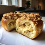 A close-up of an apple crumble cheesecake bun, sliced in half to reveal a creamy cheesecake filling and apple pieces, topped with crumble.