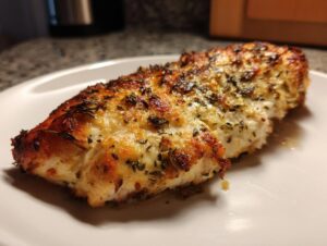 A close-up of a golden-brown Asiago chicken bake, seasoned with herbs, served on a white plate.