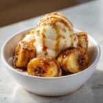 A white bowl filled with caramelized Bananas Foster pieces topped with vanilla ice cream and caramel sauce.
