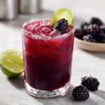 A vibrant blackberry margarita cocktail in a glass, garnished with a lime slice and fresh blackberry, with fresh blackberries and lime halves in the background.