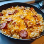 Close-up of cheesy sausage and rice cooked together in a black skillet, topped with melted cheddar cheese.