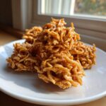A stack of golden brown, sticky haystack cookies resting on a white plate near a window.