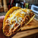 Close-up of a hand holding a crispy chicken taco overflowing with shredded chicken and melted cheese.