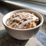 Close-up of a white bowl filled with cinnamon-spiced overnight oats topped with a thick white icing drizzle.