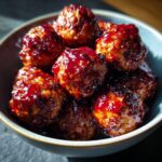 A close-up shot of glistening cranberry meatballs in a blue bowl, coated in a rich, glossy sauce.