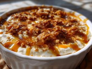 A close-up of a white dish filled with creamy cream cheese caramel apple dip, topped with crunchy bits and drizzled caramel.