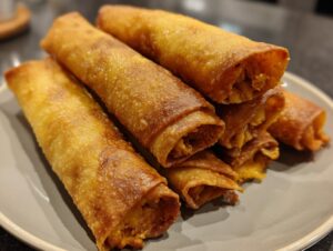 A close-up shot of a stack of golden-brown, crispy cream cheese chicken taquitos on a grey plate.
