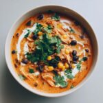 A close-up overhead view of a bowl of creamy chicken tortilla soup, topped with shredded chicken, black beans, corn, cilantro, and a swirl of cream.