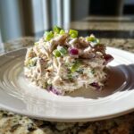 A mound of creamy crab salad mixed with red onion and celery, topped with sliced green onions, served on a white plate.