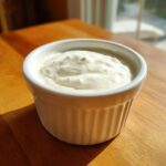 A close-up of creamy, white homemade horseradish sauce served in a small white fluted ramekin on a wooden table.