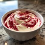 Close-up of creamy strawberry cheesecake dip swirled with bright red fruit sauce in a white bowl.