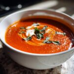 A close-up of a white bowl filled with vibrant creamy tomato soup, swirled with cream and garnished with fresh basil leaves.