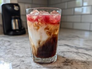 A tall glass filled with a layered Dirty Soda, featuring dark soda, creamy swirls, ice, and a red syrup topping.