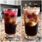A tall glass filled with a dirty soda, featuring layers of dark soda, cream, raspberries, and ice.