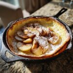 A golden-brown Dutch baby pancake with apples, dusted with powdered sugar, served in a cast-iron skillet.
