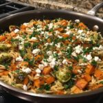 A close-up of a skillet filled with a vibrant fall orzo salad, featuring roasted butternut squash, Brussels sprouts, and crumbled feta cheese, topped with fresh parsley.