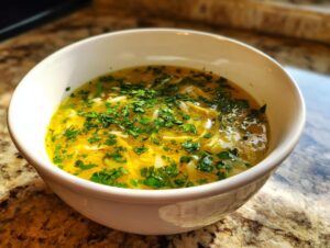 A close-up of a white bowl filled with bright yellow pastina soup topped generously with fresh, chopped parsley.