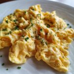 A close-up of perfectly fluffy scrambled eggs, seasoned with black pepper and garnished with fresh chives.